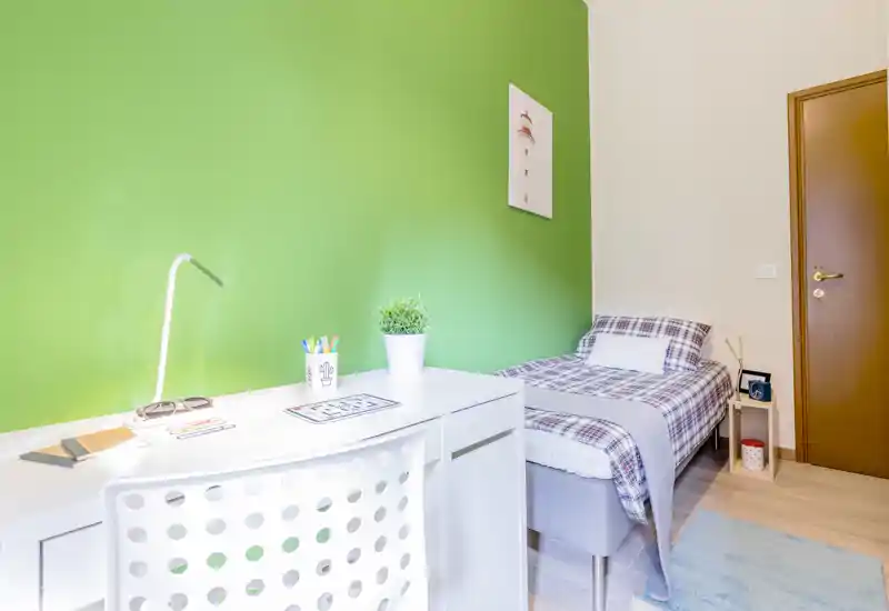 Camera da letto singola luminosa e ordinata con scrivania davanti a una parete verde vivace. Il letto è rifinito con copriletto a quadri, è presente una lampada da tavolo e un morbido tappeto: perfetta per studenti o soggiorni brevi.