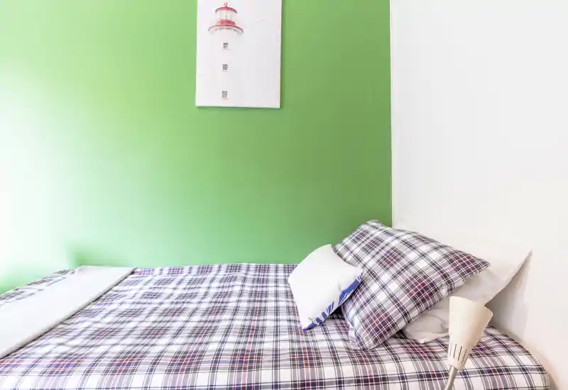 Camera da letto luminosa e ordinata con letto matrimoniale con copriletto a quadri, parete accent verde e lampada da comodino semplice — perfetta per annunci accoglienti e moderni.
