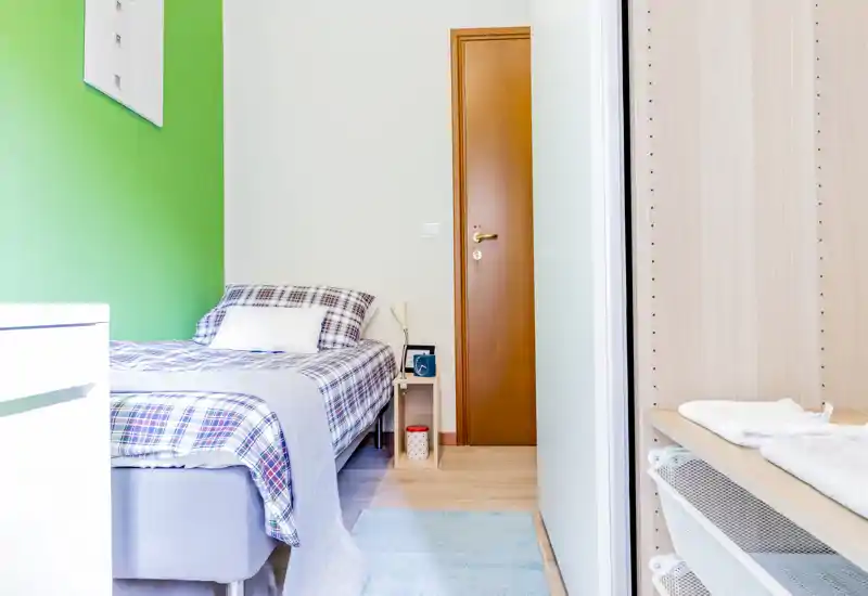 Camera da letto luminosa e arredata in modo semplice con un letto singolo coperto da una trapunta a quadri, un piccolo comodino con lampada e un armadio a muro vicino all'ingresso. Stile pulito e luce naturale gradevole, perfetta per una persona.
