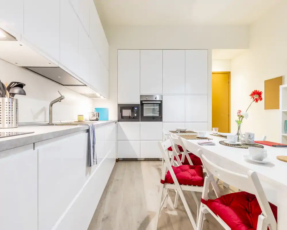 Cucina moderna e luminosa con pensili bianchi, elettrodomestici integrati e tavolo da pranzo con sedie con cuscini rossi — elegante e pronta all'uso.