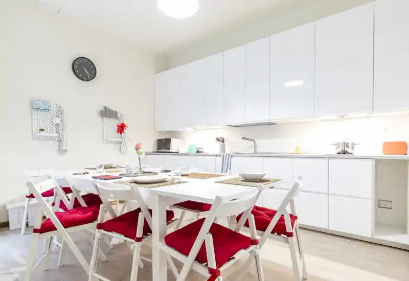 Acogedor comedor abierto y luminoso integrado en una cocina blanca. Mesa blanca amplia con sillas plegables y cojines rojos, con la mesa preparada para comidas y desayunos.