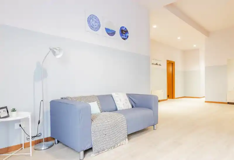 Área de estar moderna y luminosa con sofá azul de dos plazas, cojines decorativos y lámpara de pie — aireada, minimalista y bien iluminada.