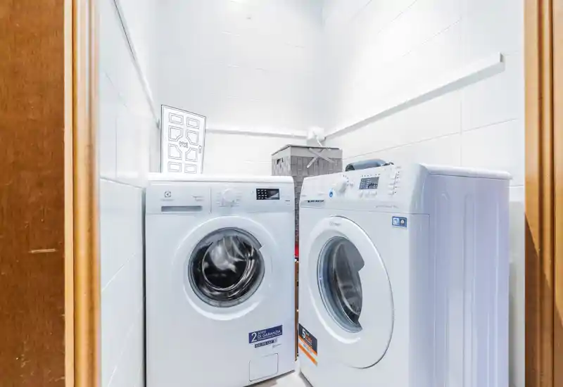 Lavadero/servicio compacto y luminoso con dos lavadoras de carga frontal, paredes alicatadas blancas y una cesta de almacenamiento — perfecto para la colada en un piso pequeño.