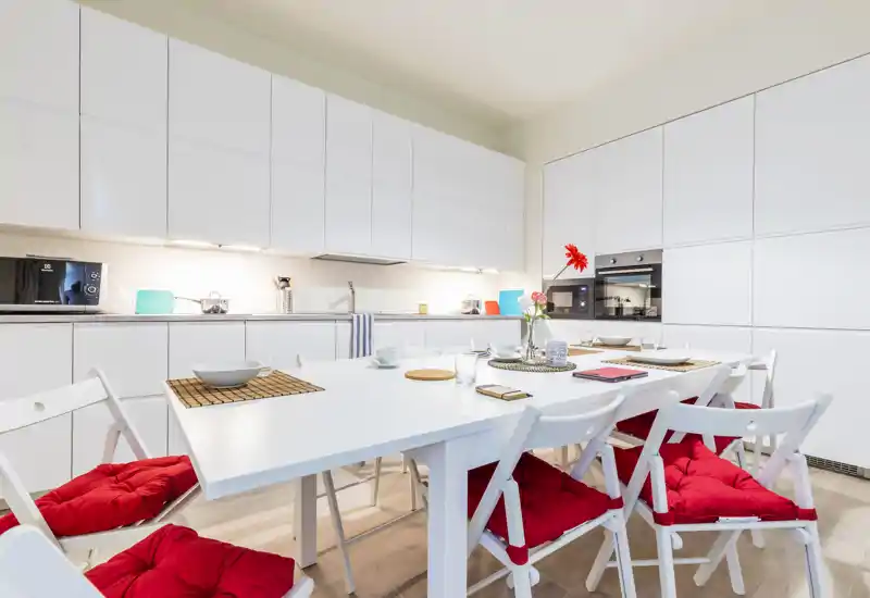 Cocina abierta moderna y luminosa con una gran mesa de comedor blanca preparada para comidas; cojines rojos en las sillas añaden color y calidez. Mobiliario blanco y electrodomésticos integrados ofrecen un aspecto limpio y contemporáneo ideal para publicar el anuncio.