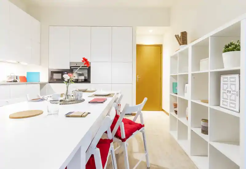 Cocina moderna y luminosa con una larga isla blanca y taburetes con cojines rojos, electrodomésticos empotrados y estanterías abiertas: espacio elegante y bien iluminado, ideal para comer o trabajar desde casa.