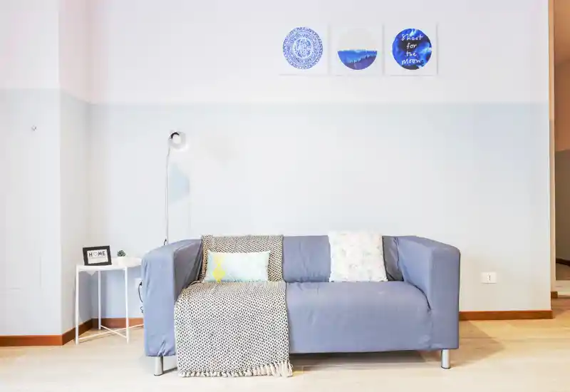 Sala de estar luminosa y minimalista con un sofá azul de dos plazas, cojines y una manta, lámpara de pie y mesita auxiliar — estilo limpio y moderno, perfecto para anuncios de alquiler.