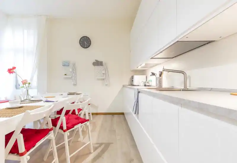 Cocina moderna y luminosa con armarios blancos integrados, fregadero y una pequeña mesa con sillas con cojines rojos — zona de cocina/comedor limpia y acogedora.