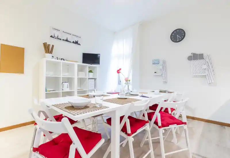 Zona de comedor moderna y luminosa con mesa blanca para varios comensales, cojines rojos en las sillas y detalles decorativos sencillos que crean un ambiente acogedor.