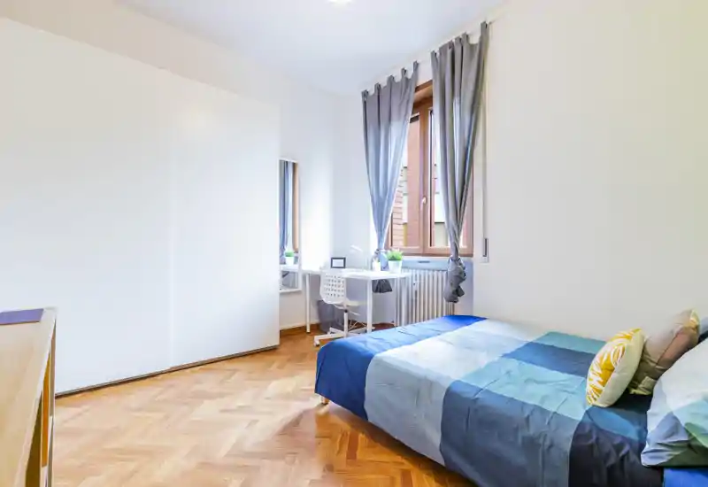 Bright, neatly styled bedroom with a double bed dressed in blue-toned linens, a tall white wardrobe and a small desk by the window letting in natural light.