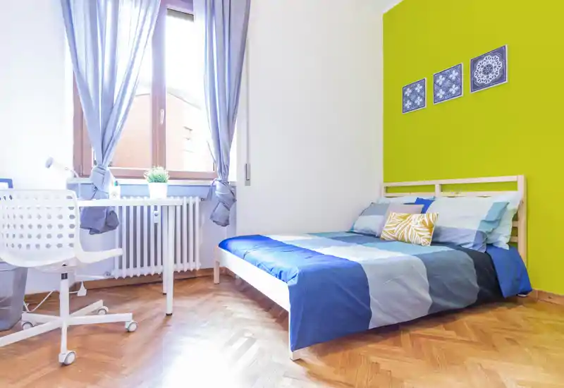 Bright, well-staged bedroom with a double bed, colorful bedding and a workspace by the window. Natural light and wooden parquet create a fresh, welcoming atmosphere ideal for rental photos.