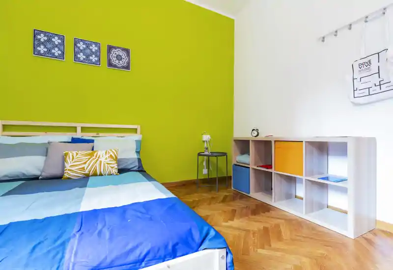Bright, modern bedroom with a blue geometric bedcover, decorative cushions and a vivid green accent wall; wooden parquet flooring and open shelving add functional storage.