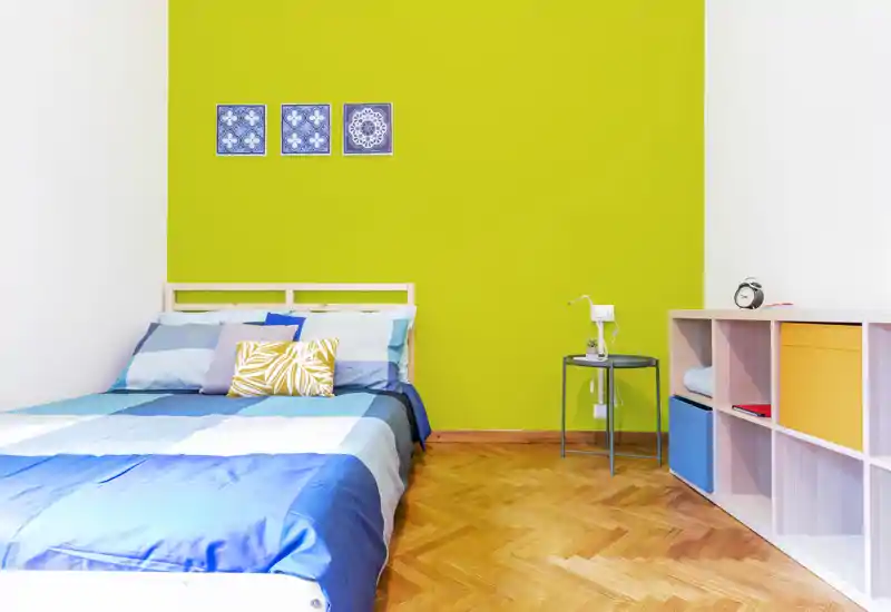 Bright, modern single/double bedroom with a colorful green accent wall, blue bed linens and neat shelving — ideal for a cheerful rental listing.