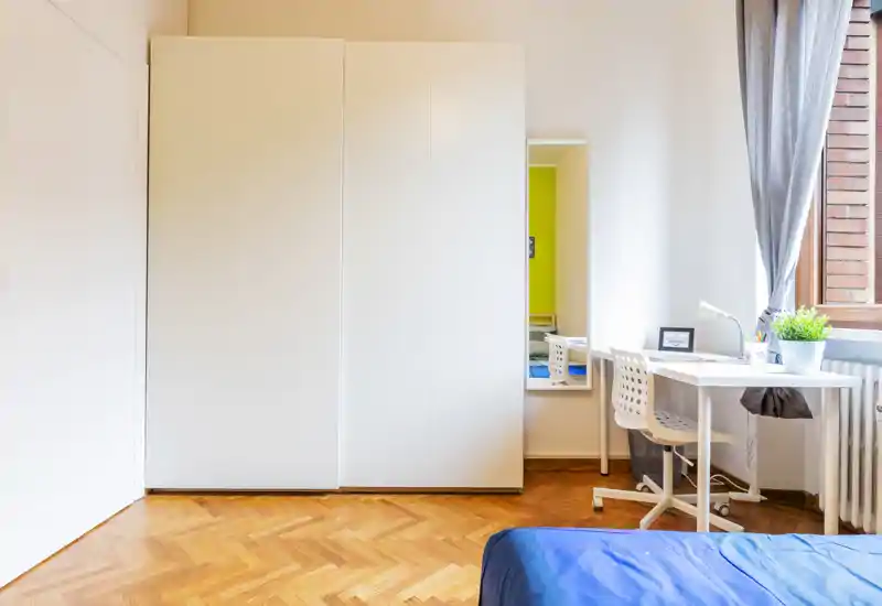 Bright, modern bedroom corner with a partially visible bed, a large white wardrobe, and a tidy desk by the window letting in natural light.