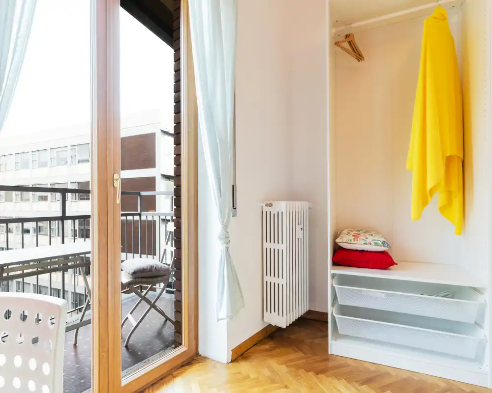 Bright corner of a living area with a door to a small balcony, parquet floor, open shelving/wardrobe and a radiator; simple, airy and well lit.