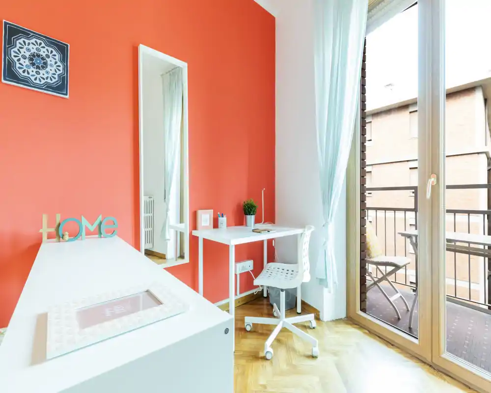 Bright, modern living/work corner with a bold coral accent wall, compact desk, chair and a balcony letting in abundant natural light.