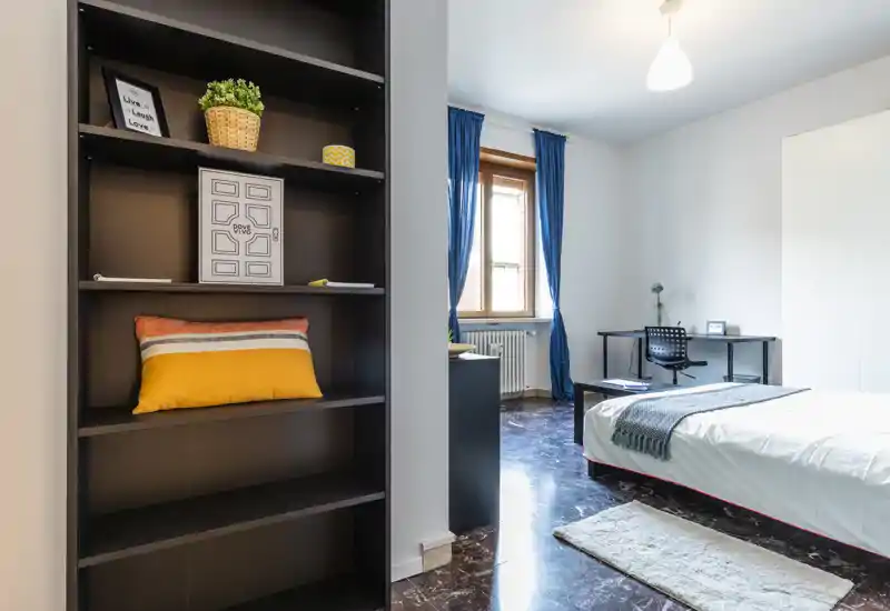 Bright, modern bedroom with a comfortable single/double bed, a desk by the window and tasteful decorative touches. Large window with blue curtains provides natural light and a neat, functional layout ideal for work and rest.