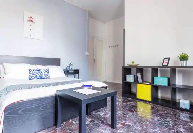 Bright, modern bedroom with a large bed, decorative cushions, bedside table and low black shelving; marble-look floor and soft natural light create a fresh, stylish atmosphere.