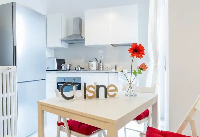 Bright modern kitchen with white cabinetry, stainless appliances and a cozy dining table set with red cushions and fresh flowers.