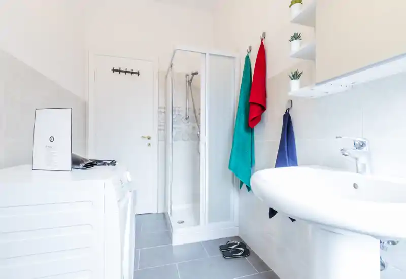 Bright, modern bathroom with a corner shower, pedestal sink and washing machine; clean white tiles and colorful towels add a fresh touch.
