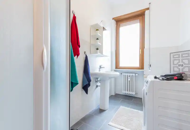 Bright, modern bathroom with a pedestal sink, natural light from a window and a washer visible — clean tiled floor and hanging towels.
