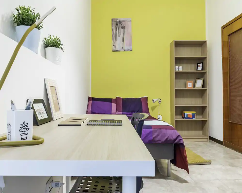 Bright, stylish single bedroom with a workspace: colorful bedding against a yellow accent wall, a practical desk in the foreground and a tall shelving unit for storage.