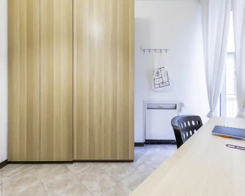 Bright, minimal bedroom corner with a large wooden wardrobe, desk edge and sheer curtains letting in natural light; clean tiled floor and simple decor create a calm, modern feel.