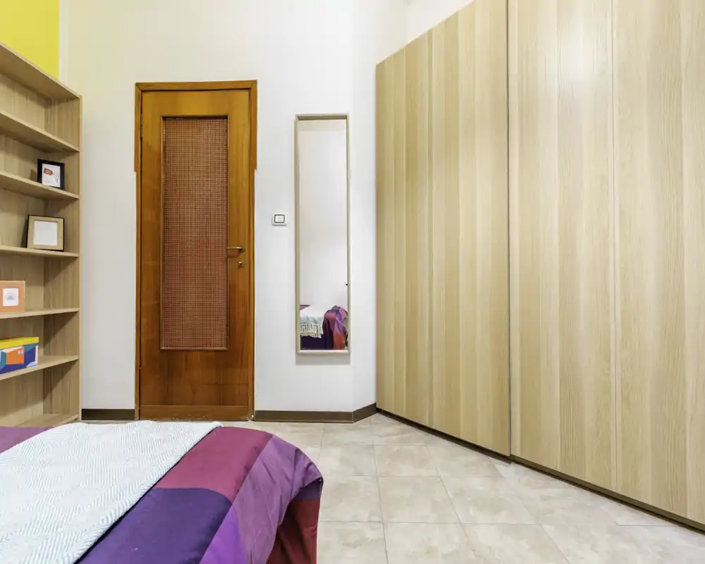 Bright, tidy bedroom corner with a colorful bedspread in the foreground, a full-height wardrobe and wall mirror near the wooden door.