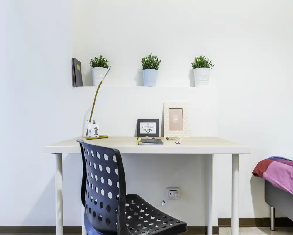 Bright, minimal bedroom corner with a simple desk and chair; decorative plants and framed prints add a fresh touch. A small portion of the bed is visible at the right edge, indicating this is a bedroom workspace.