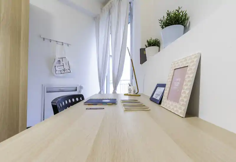 Bright, minimal workspace in a living area: a long wooden desk with decorative frames, plants and stationery by a sunlit window.