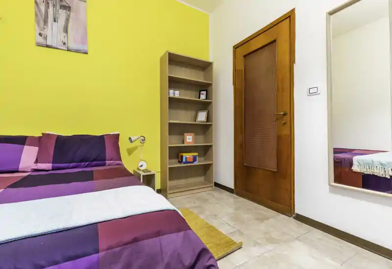 Bright, colorful bedroom with a double bed dressed in geometric purple and magenta bedding, a bedside lamp, open shelving and a full-length mirror — ideal for a cheerful rental listing.