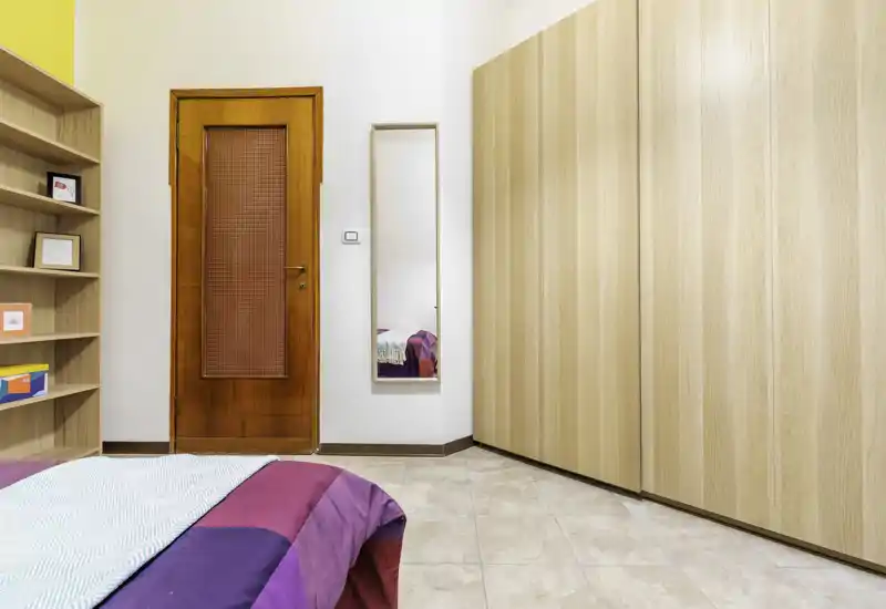 Bright, tidy bedroom corner with a colorful bedspread in the foreground, a full-height wardrobe and wall mirror near the wooden door.