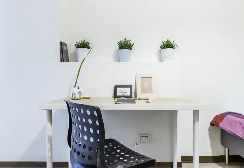 Bright, minimal bedroom corner with a simple desk and chair; decorative plants and framed prints add a fresh touch. A small portion of the bed is visible at the right edge, indicating this is a bedroom workspace.