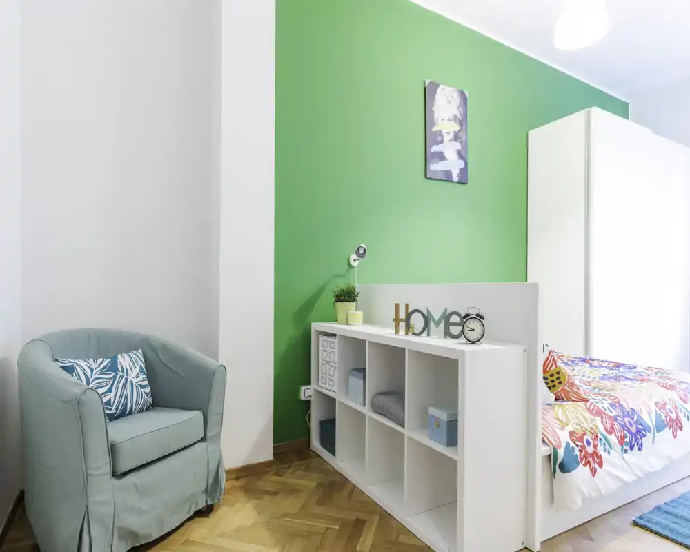 Dormitorio luminoso y colorido con edredón estampado, estantería blanca y butaca acogedora: espacio moderno y bien decorado, ideal para una persona.