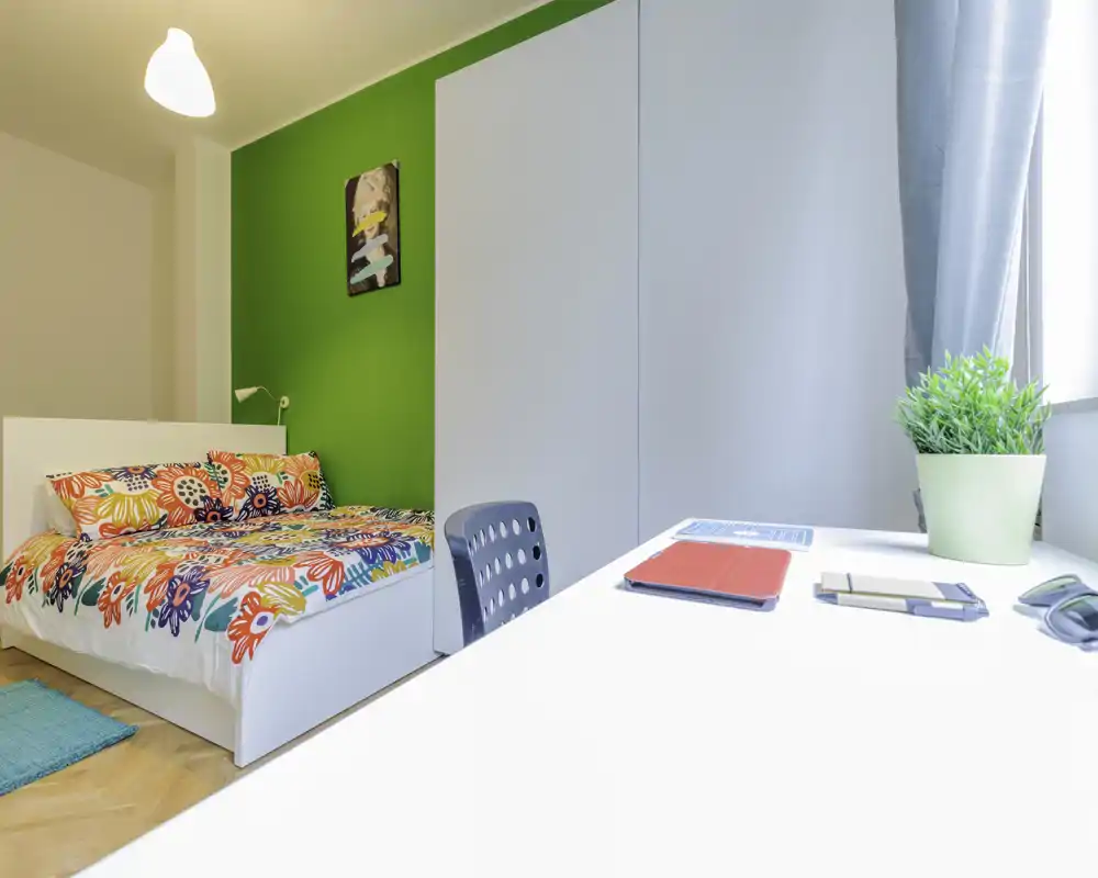Dormitorio luminoso y colorido con cama doble, ropa de cama floral, escritorio junto a la ventana y gran armario — ideal para estudiante o persona sola.