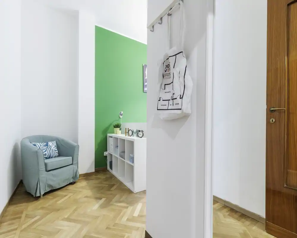 Zona de estar compacta y luminosa con un sillón cómodo, cojín decorativo y estantería baja frente a una pared verde llamativa.