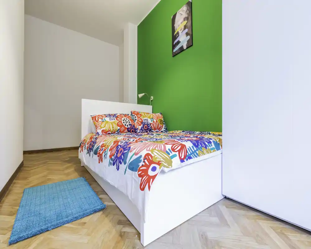 Dormitorio luminoso y moderno con edredón floral colorido, cama blanca y pared verde intensa. Suelo de parquet en espiga y alfombra turquesa que completan un estilo fresco y cuidado.