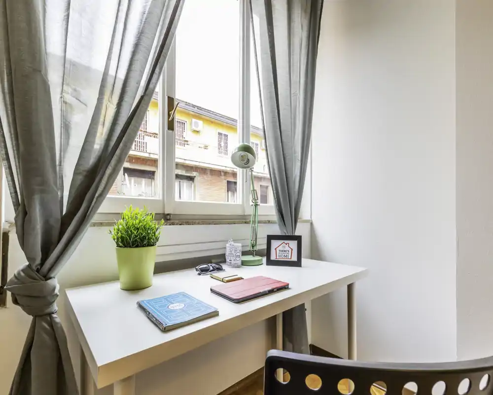 Espacio de trabajo luminoso junto a una gran ventana con escritorio blanco, lámpara, planta en maceta y material ordenado — un rincón de oficina en casa compacto y acogedor.