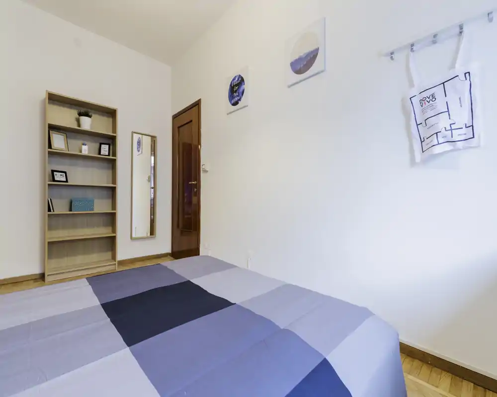 Camera da letto moderna e luminosa con letto matrimoniale ben rifatto in toni blu-grigio, libreria in legno, specchio a figura intera e decorazioni murali semplici — perfetta per un annuncio essenziale.