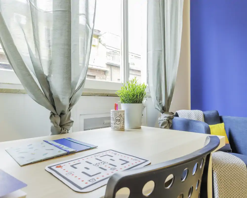 Zona giorno luminosa e accogliente con tavolo in legno vicino a una grande finestra, tende leggere, una pianta in vaso e un comodo divano blu con cuscini colorati.