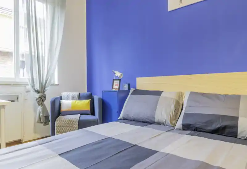 Camera da letto moderna e luminosa con letto matrimoniale, biancheria a quadri, testiera in legno e una poltrona accogliente vicino alla finestra. La parete blu vivace e la luce naturale creano un ambiente fresco e invitante, ideale per affitti brevi.