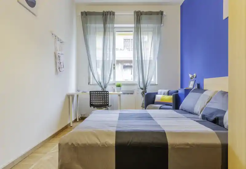 Camera da letto luminosa e moderna con un ampio letto matrimoniale, scrivania vicino alla finestra e una poltrona accogliente, ideale per rilassarsi o lavorare.