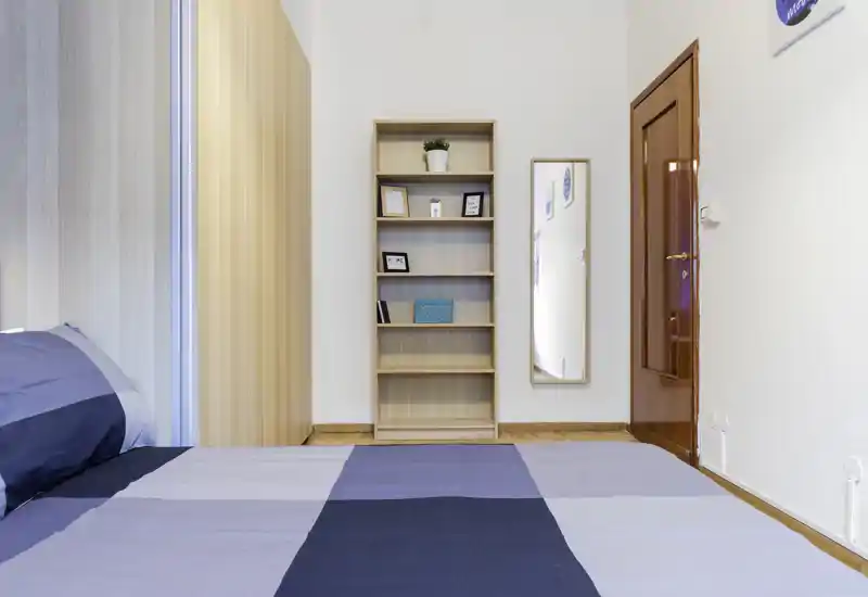 Camera da letto luminosa e minimal: letto ampio in primo piano, scaffale in legno, specchio a parete e porta in legno — perfetta per un annuncio moderno e pulito.