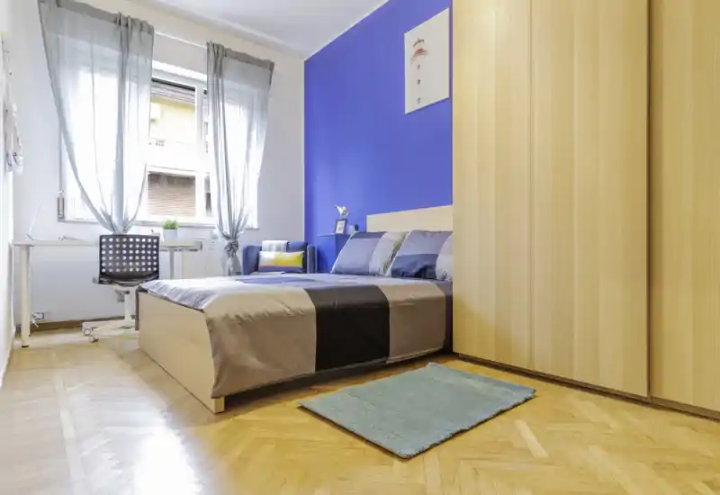 Camera da letto luminosa e ben arredata con letto matrimoniale contro una parete blu, armadio in legno e scrivania vicino alla finestra — ideale per un soggiorno confortevole.