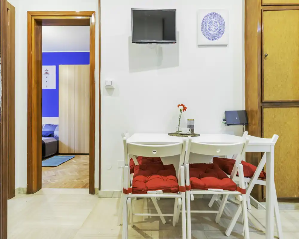 Angolo pranzo compatto con tavolo bianco e sedie pieghevoli con cuscini rossi, pareti luminose e un piccolo centrotavola decorativo—ideale per pranzi intimi da due a quattro persone.
