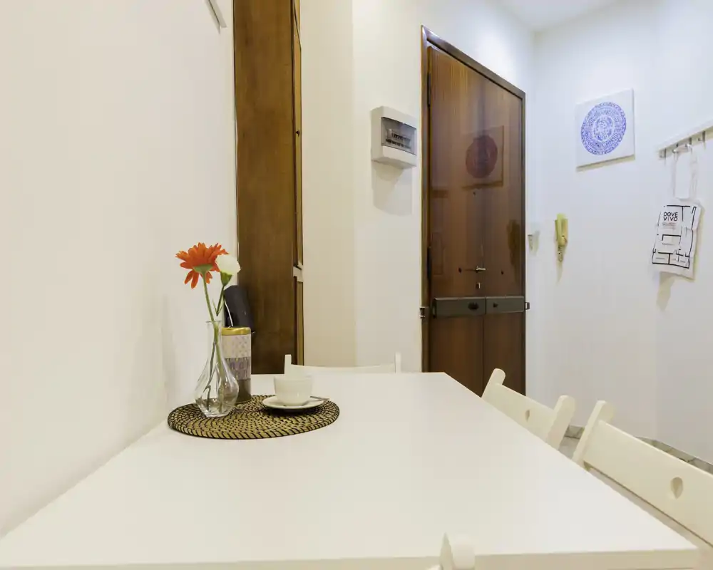 Zona pranzo luminosa e minimale con un tavolo bianco apparecchiato per una persona, un sottopiatto in vimini e un fiore in un vaso di vetro — pulita e accogliente.