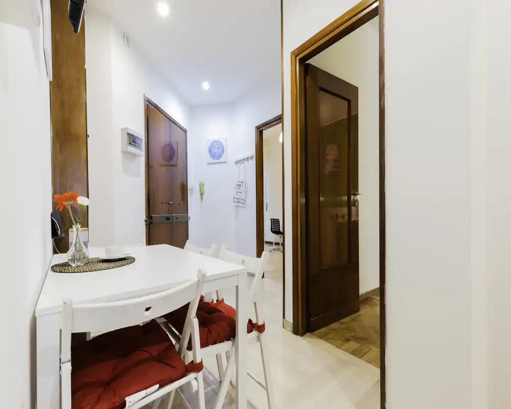 Angolo pranzo compatto vicino all’ingresso con tavolo bianco, sedie pieghevoli con cuscini e semplici dettagli decorativi — luminoso e accogliente per pasti veloci.