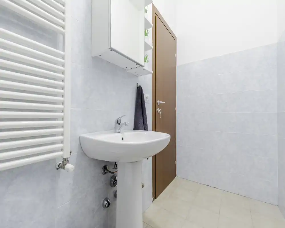 Bagno pulito e luminoso con lavabo a colonna, termoarredo a parete e porta in legno; pareti piastrellate azzurro chiaro e pavimento neutro.