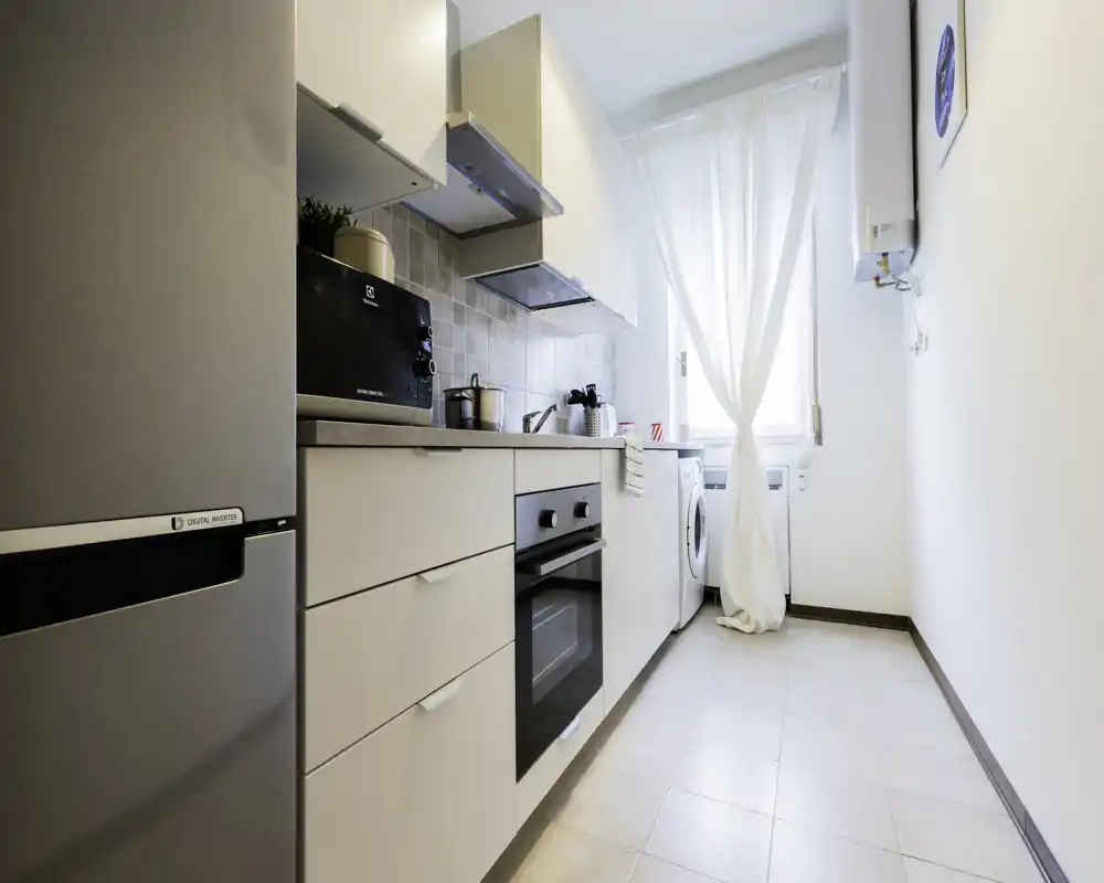 Cucina moderna e luminosa a corridoio con forno integrato, frigorifero e lavatrice. Pensili bianchi, paraschizzi piastrellato e tenda leggera per un’atmosfera fresca e ariosa.