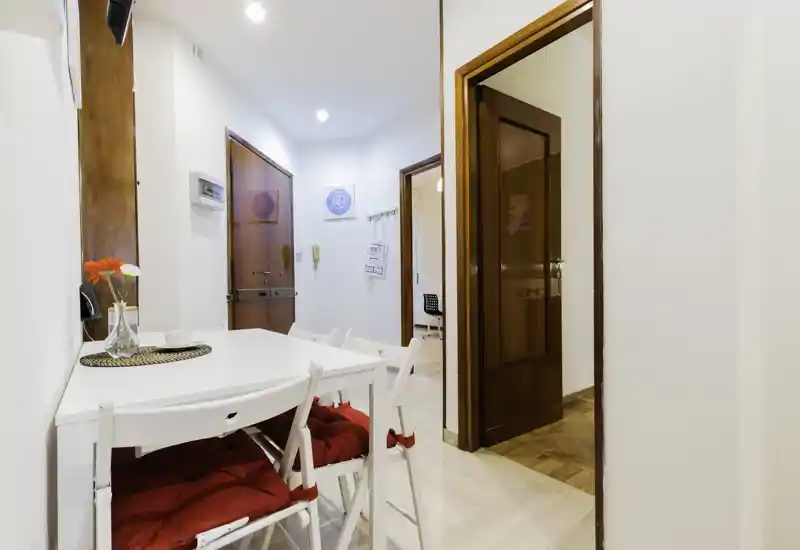 Compact dining area near the apartment entrance with a white table, cushioned folding chairs and simple decorative details — bright and welcoming for quick meals.