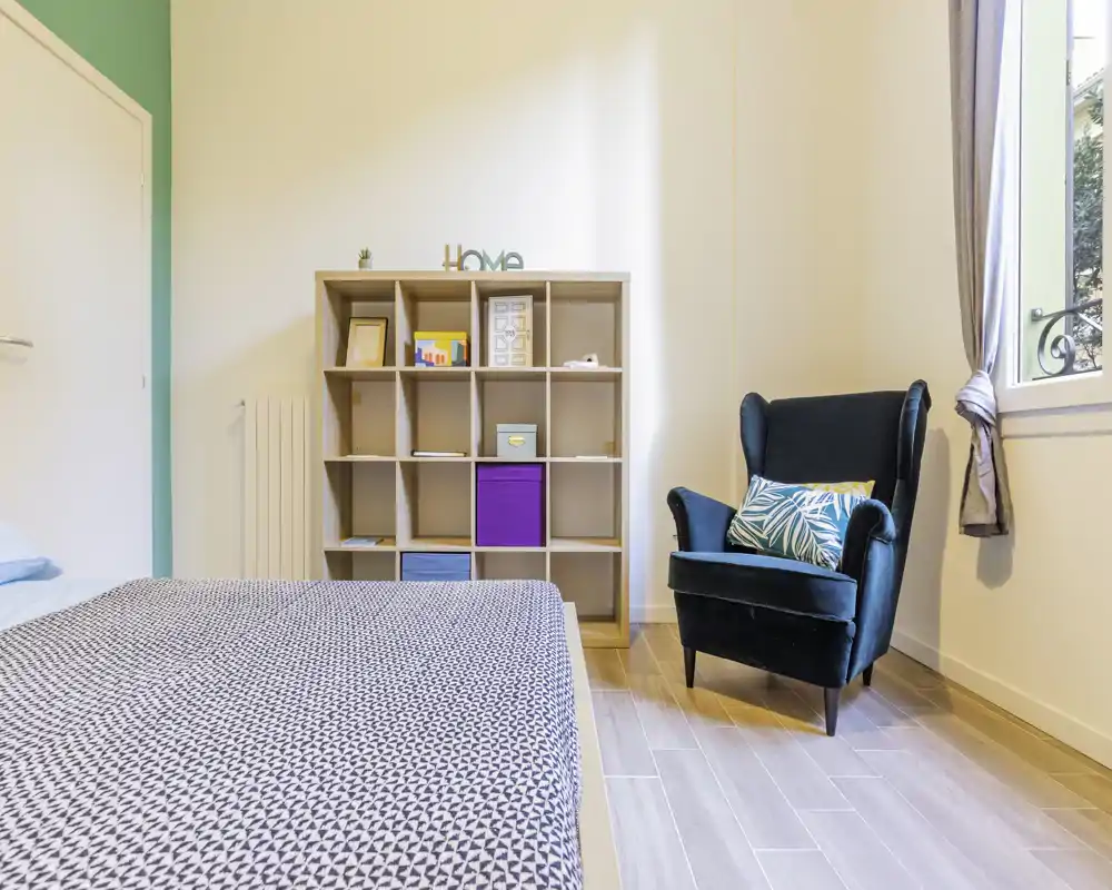 Camera da letto moderna e luminosa con copriletto a motivo, poltrona in velluto e scaffalatura aperta — luce naturale e tocchi di colore gradevoli.